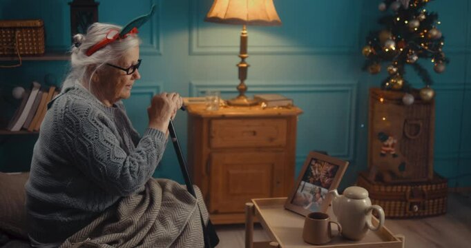 The old grandmother celebrates Christmas alone at home, feels sad and lonely sitting on the bed, and looks at family photos. Sad retired senior 90s woman who misses her family during winter holidays