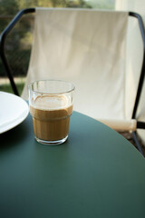 From above glass with foamy coffee placed on green table with white curtains in soft light.