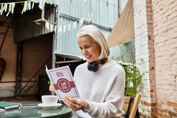 A joyful woman with gray hair sits at a cafe table, eagerly reading the menu while sipping coffee.