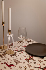 White tablecloth with red reindeer, on which there are golden candlesticks with white candles, glass glasses on slender legs and ceramic plates the color of warm chocolate.
