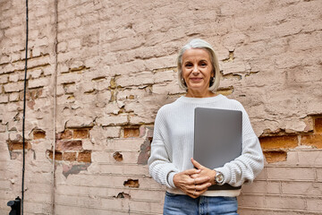 Fototapeta premium A sophisticated gray haired woman smiles warmly with her laptop against a brick backdrop.