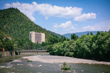 The calm River Kura flows with green tree-covered mountains in the background, framing a Soviet-era building. Captured in summer in Borjomi, Georgia, the scene reflects tranquility and natural harmony © Anna