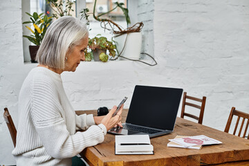 A beautiful mature woman with gray hair interacts with her smartphone at a cozy workspace.