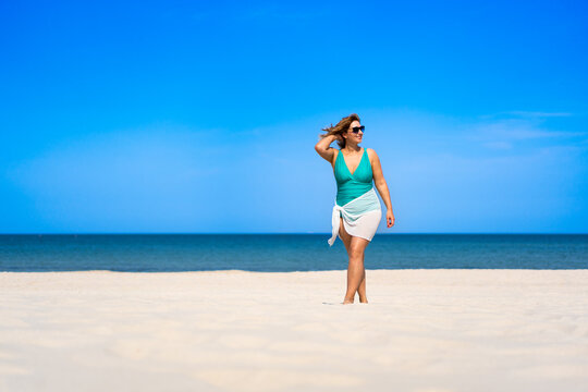 Summertime. Beautiful young woman in one-piece swimsuit and white sarong and sunglasses walking on white sandy beach on sunny day. Sunbathing. Front view.