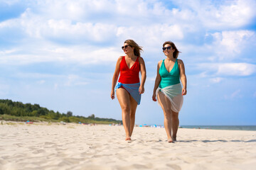 Two beautiful young females wearing one-piece bathing suits and pareo walking on white sandy beach in summertime. Front view.