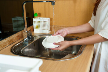 Young Asian woman in casual white shirt washing a white plate under running water at the kitchen sink, focused on cleaning with attention to hygiene and cleanliness in a simple indoor kitchen setting.