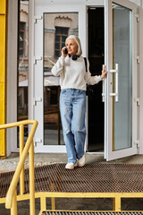 A mature woman in a cozy sweater speaks on the phone while standing at the door.