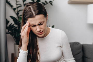 Dizzy young female with headache or migraine pain suffering from vertigo while sitting on couch in...