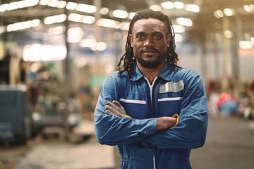 A man with dreadlocks is wearing a blue jumpsuit and is standing in a factory. He is smiling and looking directly at the camera