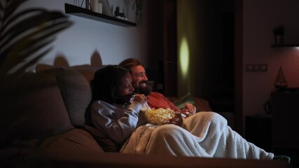 Side view of young multiracial couple relaxed watching late night movies lying on sofa at home eating popcorn. Two people together covered with a blanket in a dark living room enjoying a tv serie. - Powered by Adobe