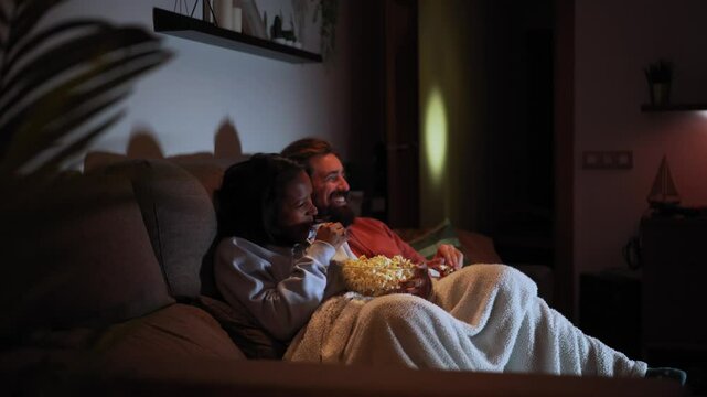 Side view of young multiracial couple relaxed watching late night movies lying on sofa at home eating popcorn. Two people together covered with a blanket in a dark living room enjoying a tv serie.