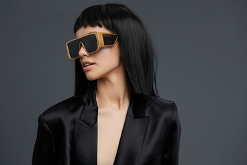 Woman with black hair, sunglasses, and a black blazer standing against a gray background with a...