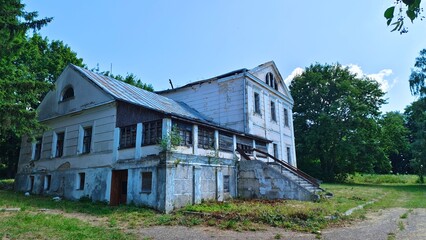 An old abandoned stone house with a metal roof and window frames without glass is surrounded by a grassy lawn and trees. A paved road approaches the front porch with a railing. Sunny summer weather