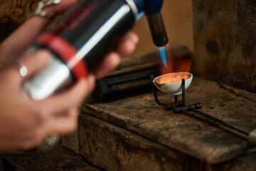 A skilled artisan uses a blowtorch to melt metal, creating exquisite jewels in a cozy workshop.