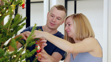 Young couple is adorning their home's christmas tree with handcrafted glass ornaments, preparing...