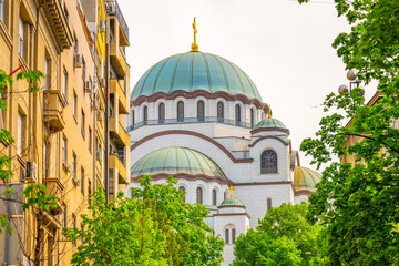 Saint Sava Cathedral stands majestically against a summer sky, surrounded by lush greenery and historical buildings. Visitors enjoy the vibrant atmosphere in Belgrade.