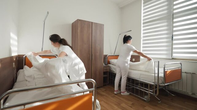 Two healthcare workers change bed linens in a brightly lit hospital room, ensuring cleanliness and preparation for patient care. Concept of hygiene, teamwork, and healthcare standards.