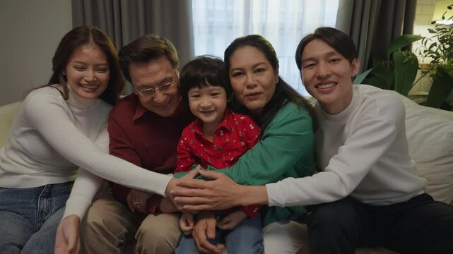 Portrait of Asian parents, child, and grandparents sitting closely and hugging on a sofa in a cozy living room, enjoying warm family moments at home.