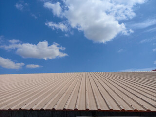 corrugated metal roof under a bright blue sky with scattered white clouds. The roof is beige in color and has evenly spaced ridges running vertically. The sky is clear with a few fluffy clouds.