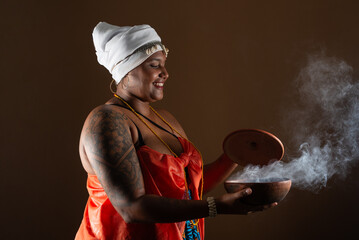 Beautiful smiling woman in traditional Candomble clothes, opening clay pot with smoke coming out of...