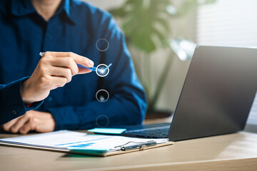 mark, checklist, list, scratch, checkmark, job, meeting, report, review, tick. A man is sitting at a desk with a laptop and a pen. He is holding the pen in his hand and looking at the laptop screen.