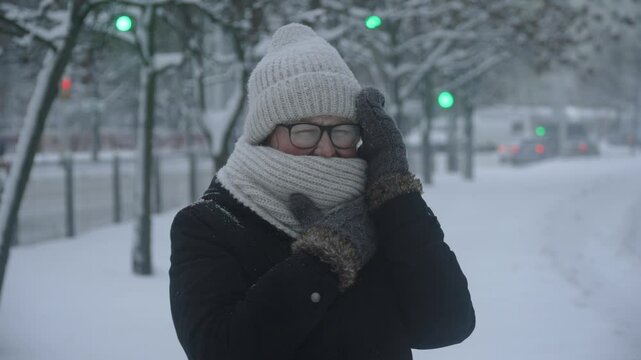 Caucasian woman with glasses wrapped in scarf trying to hide from snowfall in city. Bad weather with wind due to hurricane. Portrait of person suffering during bad weather on cold winter evening.