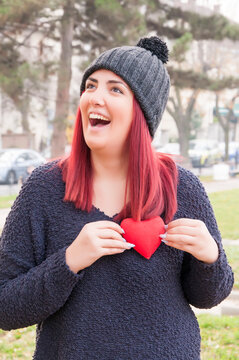 Portrait of a sedactive redhead with woolen hat in the park, holding a red heart