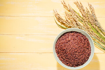 A bowl of red rice is placed on a yellow table next to ears of rice.The bowl is filled with raw rice.The rice is spread out in the bowl.copy-space.