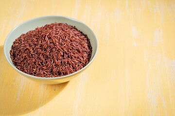 A bowl of red rice is placed on a yellow  table.The raw rice is spread out in the bowl, copy-space.