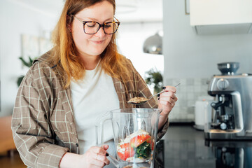 Plus size young woman adding flex seeds during making smoothie on the kitchen. Superfood supplement. Healthy detox vegan diet. Healthy dieting eating, weight loss program. Selective focus.