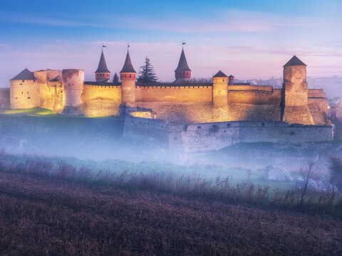 landscape view to medieval castle with towers in illumination in morning twilight with fog under castle walls