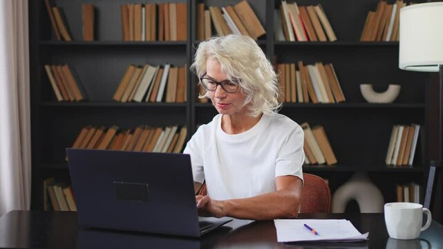 Confident stylish middle aged senior woman using laptop at workplace. Older mature lady businesswoman sitting at office table with laptop and paper document. Boss leader teacher professional worker