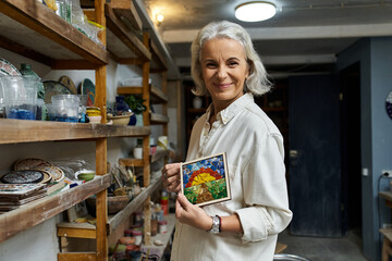 In a warm workshop, a mature woman holds up her vibrant painting, smiling with pride.