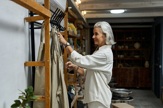 The mature woman gently arranges her garments on a wooden rack in a warmly lit space.