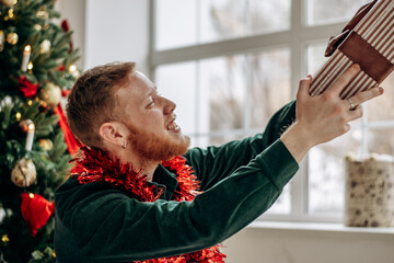 A happy red-haired young man is holding a present in his hands. Close-up on the background of a Christmas tree.New Year and Christmas concept.