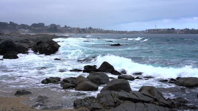 Turquoise ocean waves crash against a rocky beach, with a charming town visible in the distance along the coastline