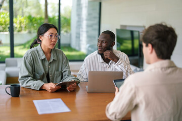 A young Asian woman and African man collaborate with a colleague in a well-lit office, surrounded by digital devices and work materials.