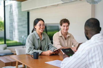 A group of joyful diverse young business professionals collaborates using a tablet in a well-lit modern office space, dressed in casual business attire.