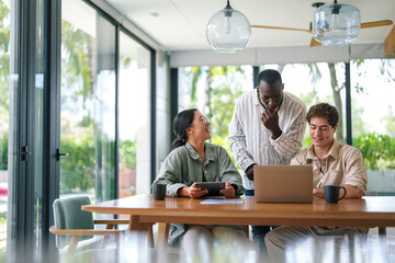 A cheerful diverse group of young adult professionals engage in a discussion over a laptop, dressed in casual business attire, in a bright and modern office environment.