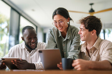 A group of three young adult professionals collaborates with enthusiasm in a sunlit office, utilizing digital tablets and sharing ideas.