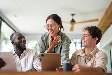 A diverse group of joyful young adults collaborates with digital tablets and laptops in a modern, bright office, wearing casual professional attire.