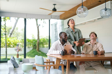 A diverse group of young adult business professionals engage in a lively discussion at a wooden table, with digital tablets and coffee cups in hand, in a spacious and sunlit modern office.