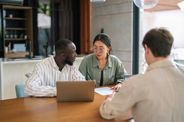 A young Black male and Asian female professional engage in a focused discussion over a laptop, alongside a colleague, in a sleek, contemporary workspace.