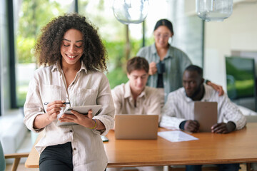 A cheerful African American young adult businesswoman interacts with a digital tablet. Colleagues work in the background of the well-lit, contemporary office space.
