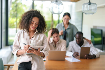 African American young adult businesswoman appears engaged and joyful using a digital tablet. She's in a modern office, dressed smartly with diverse colleagues in the background.