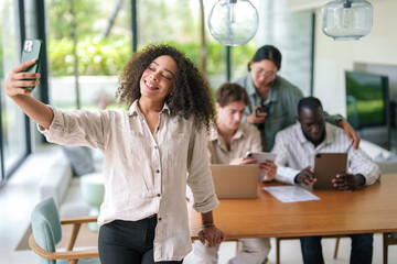 Obraz premium An African American businesswoman enjoys a light moment, capturing a selfie with a smartphone, while her diverse colleagues work in the background of a bright, contemporary office.