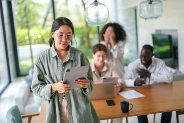 A content Asian young adult businesswoman uses a digital tablet, engaging in work with a diverse team in a bright, contemporary office environment.