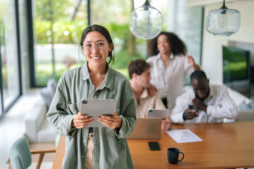 A joyful Asian young adult businesswoman interacts with a digital tablet, her attire casual professional, in a lively diverse office setting with colleagues.