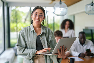 A joyful Asian young adult businesswoman uses her laptop while standing in a brightly lit modern office, with diverse colleagues focused on work in the background.