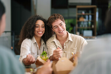 A cheerful group of diverse young business professionals enjoys a casual lunch together, engaging in conversation amid the sleek surroundings of a contemporary office space.